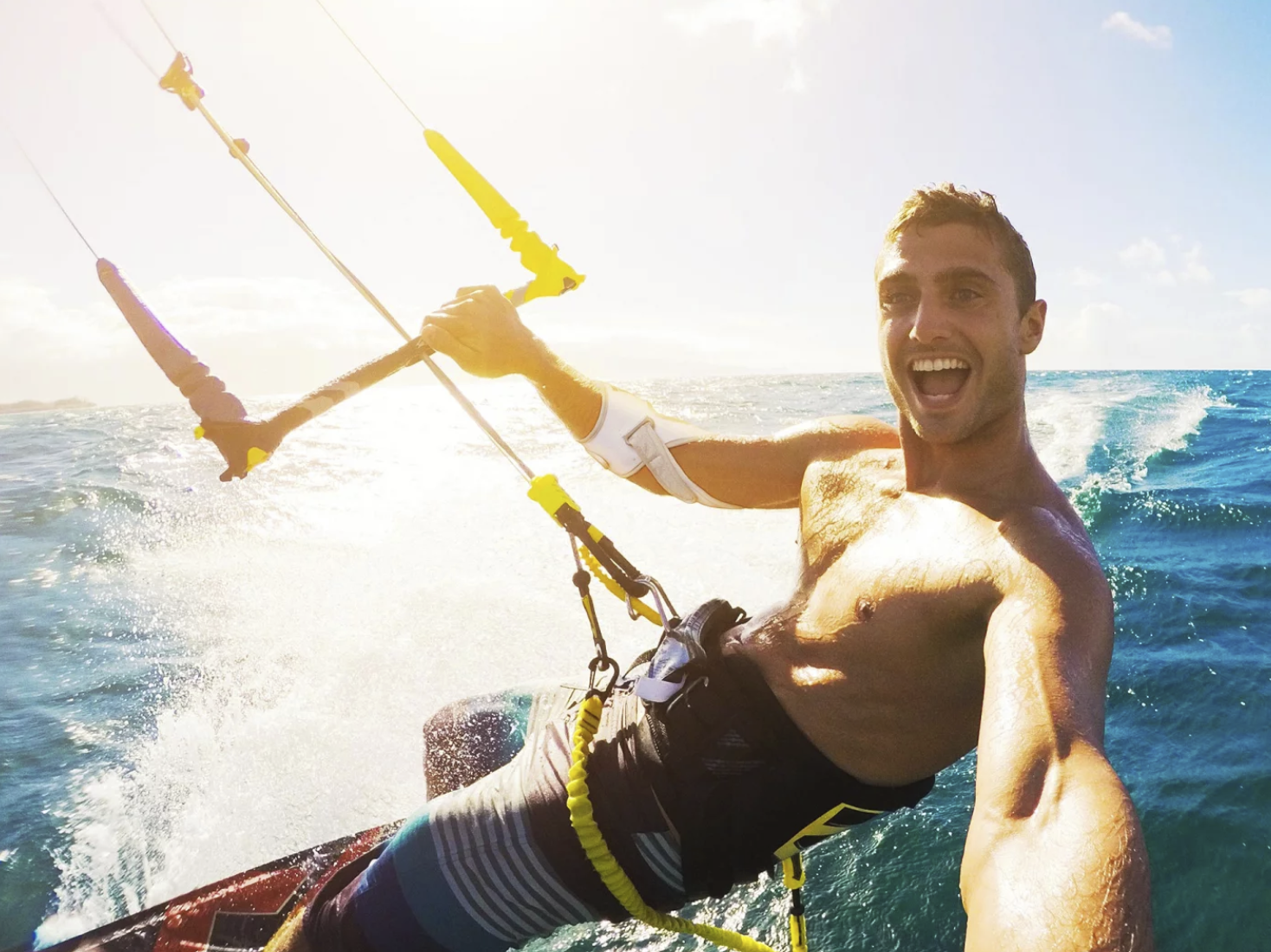 Masalo Manschette tragender junger, sportlicher, lachender Mann beim Kitesurfing im Meer.