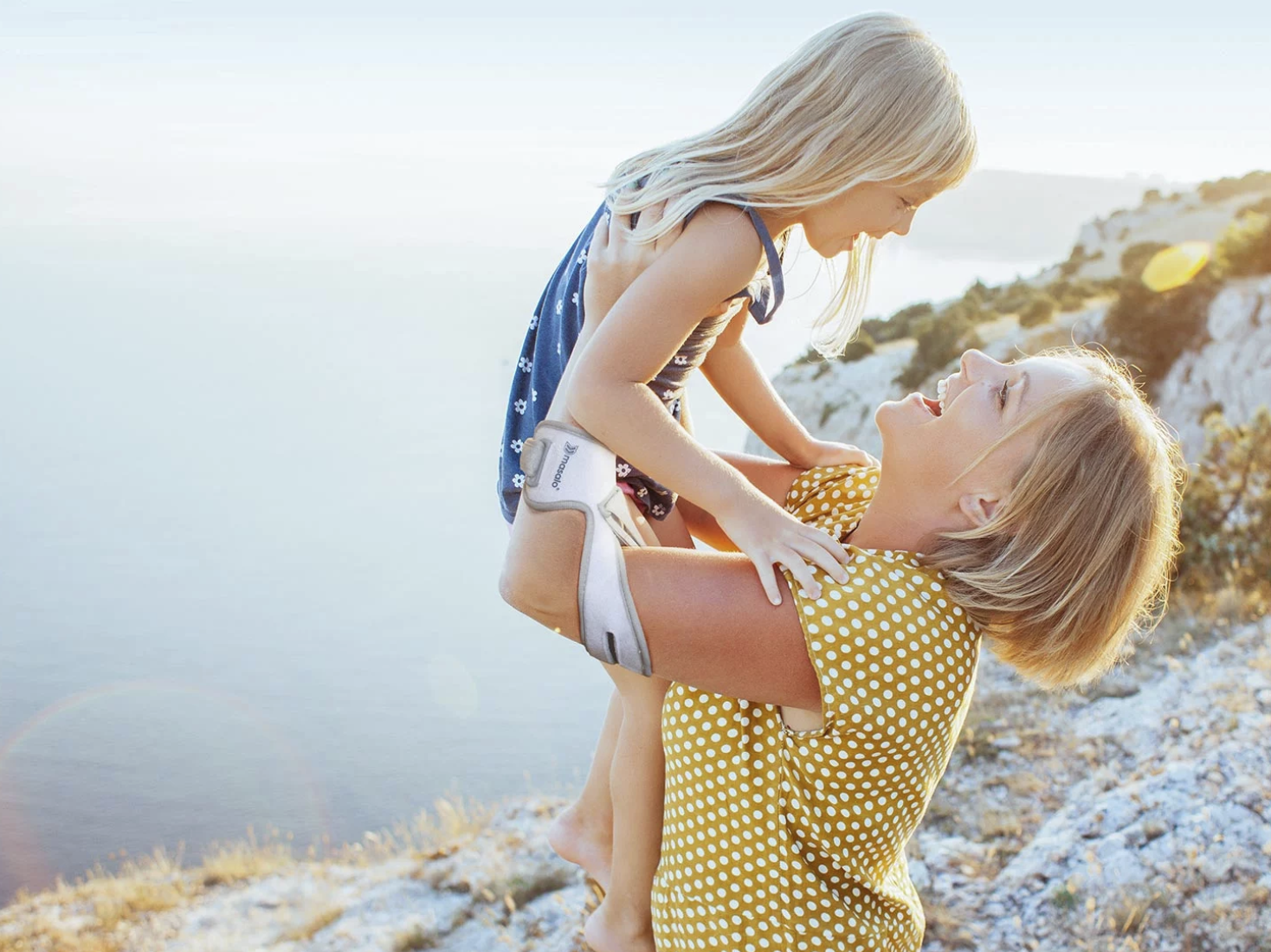 Masalo Manschette - Für mehr Lebensqualität im Alltag, Beruf & Sport Masalo Manschette tragende junge Frau hebt ihre junge Tochter in die Luft. Die beiden stehen an einer Klippe am Meer.