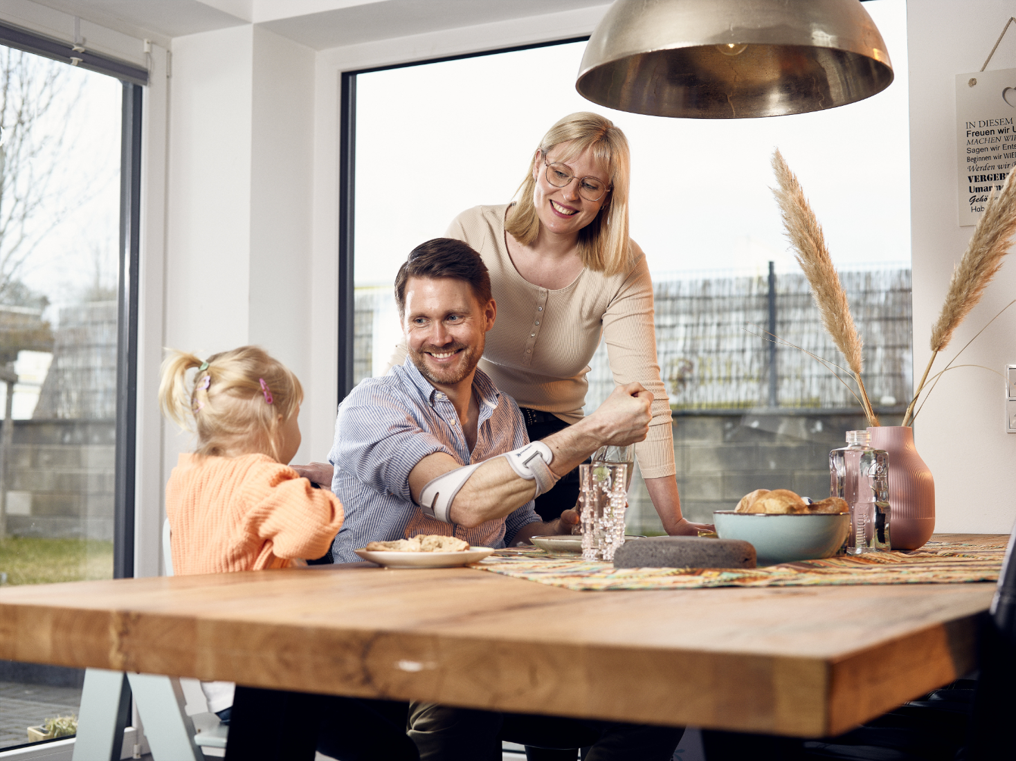 Masalo Manschette - lindert Schmerzen und unterstützt deinen Arm auch im Alltag Masalo Manschette tragender Vater sitzt am Frühstückstisch mit seiner Familie und öffnet eine Wasserflasche